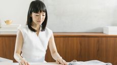 Marie Kondo in a white linen sleeveless blouse folding clothes on a table. Background shows a mid century wooden sideboard which is tidy and clear of clutter