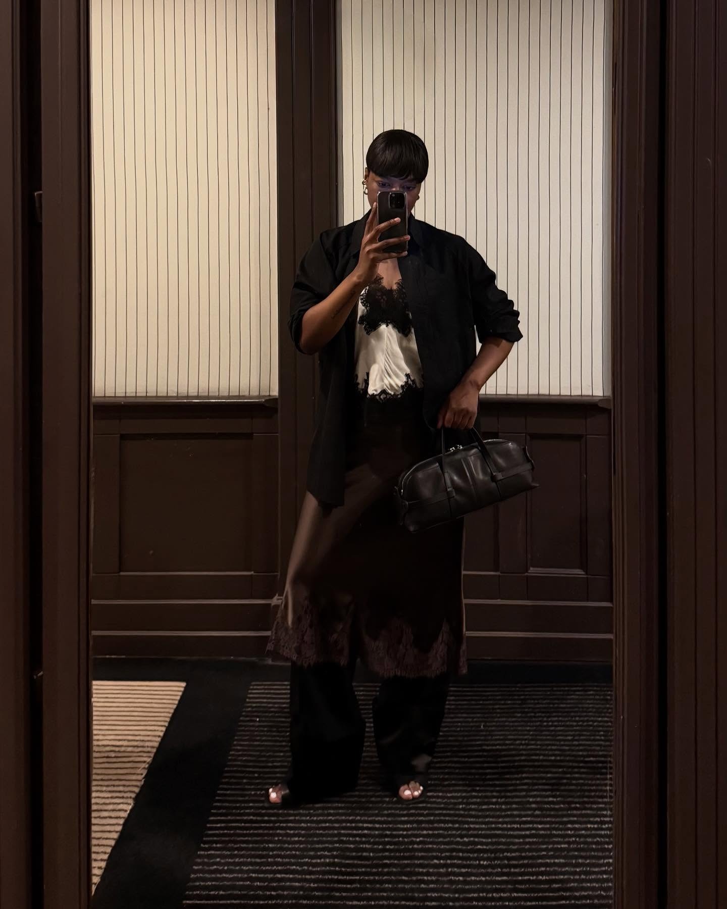 Grace wears a black jacket, white and black lace top, brown skirt, and black trousers.