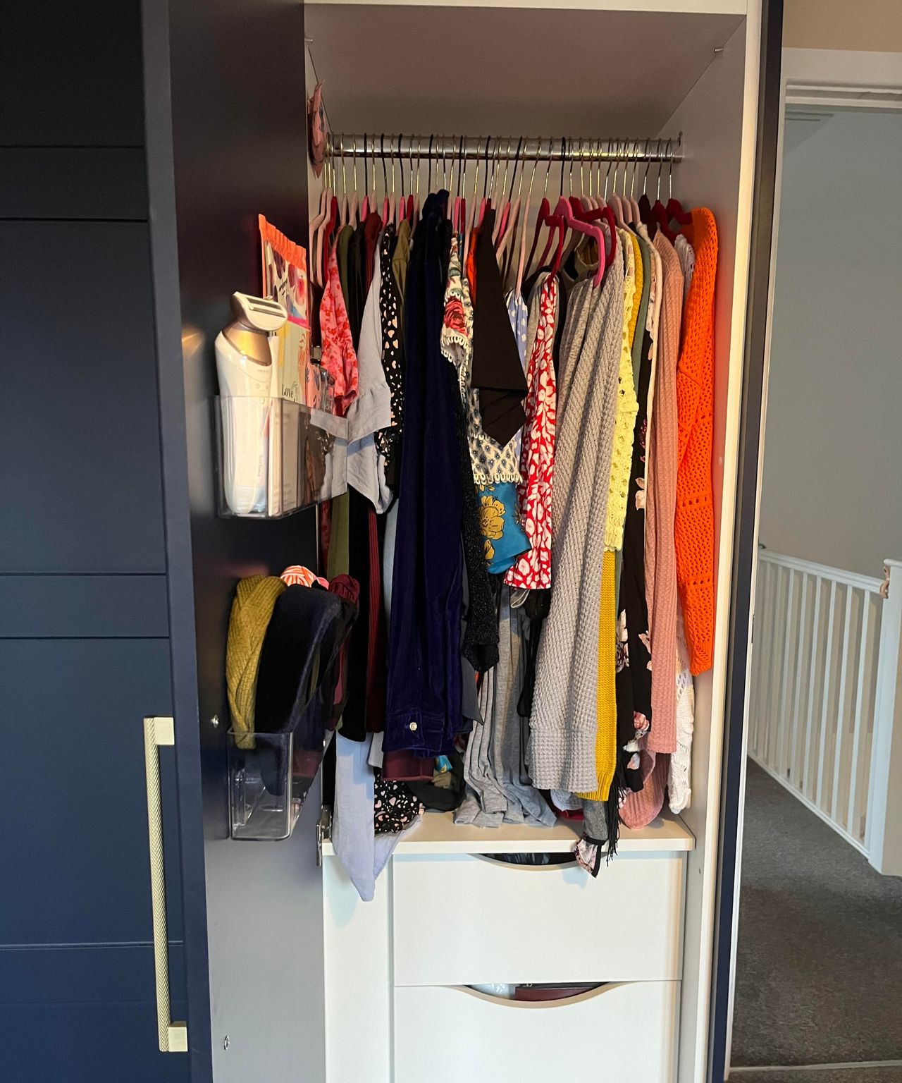 Side on view of my clear plastic adhesive organizers in my matte dark blue closet which is open and shows hanging rail of clothes directly ahead with white drawers under