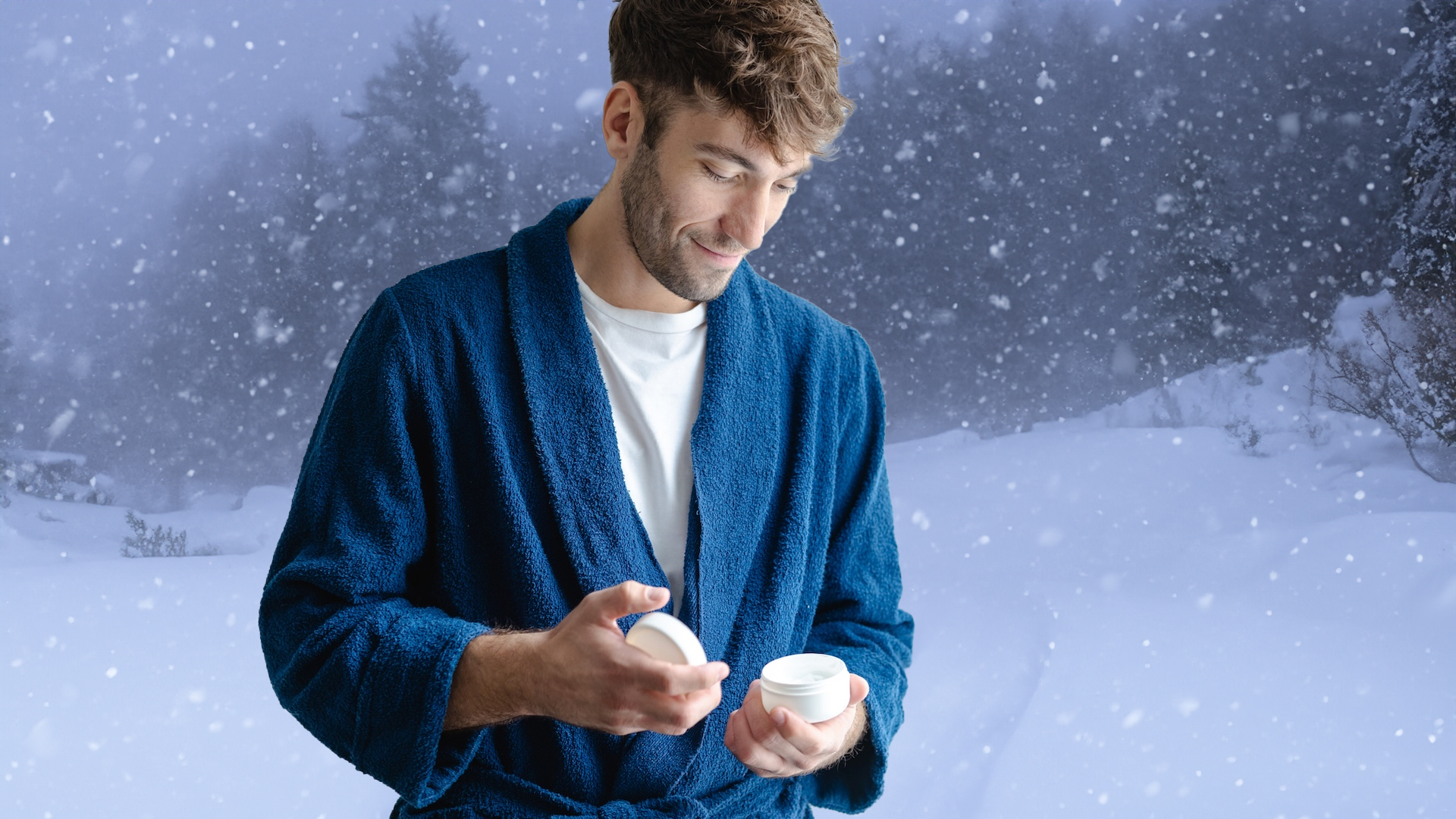 A man in a blue bathrobe applies moisturizer from a small jar in a bright, modern bathroom by a window, conveying a calm morning skincare routine and self-care at home.