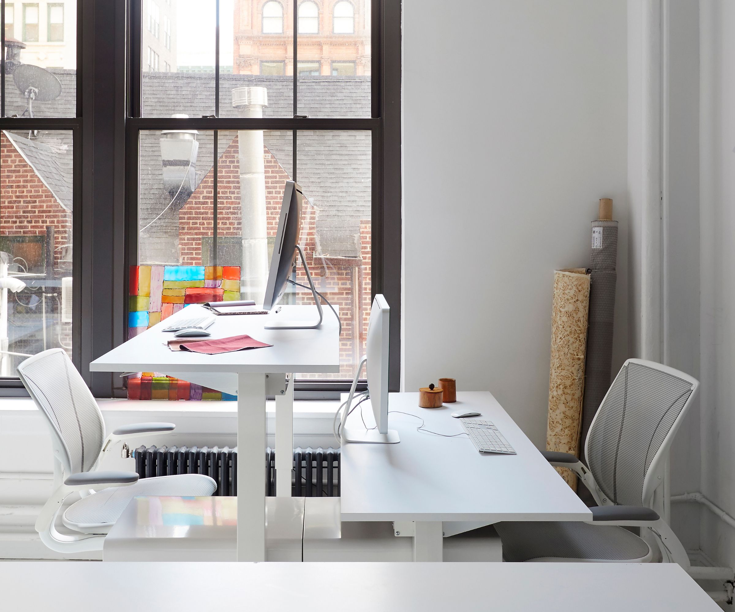 A bright, modern office with two white desks—one raised for standing, one seated—paired with mesh ergonomic chairs and large windows overlooking rooftops.
