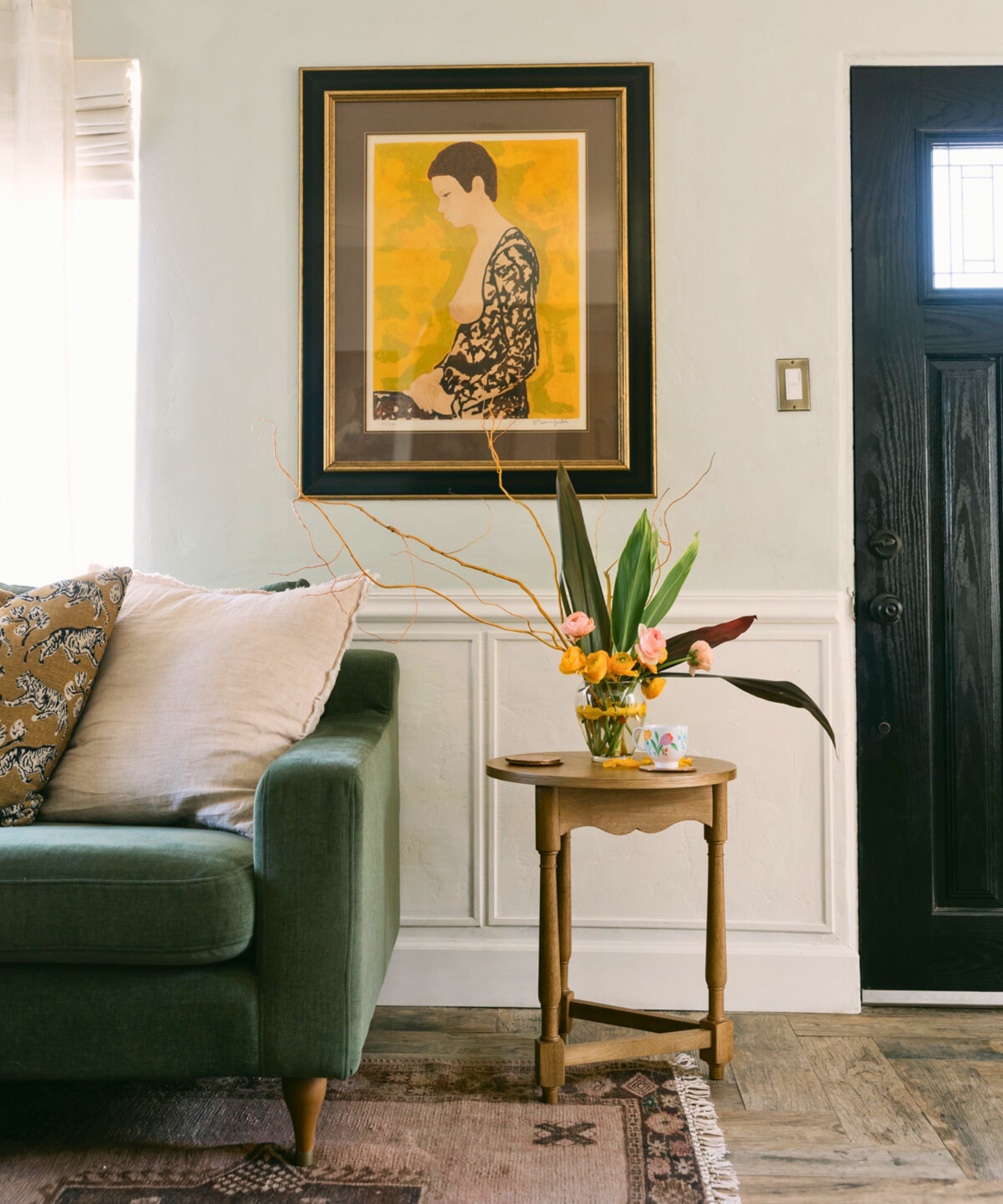 Photograph of a home's muted wall, behind a side table with plants, a framed painting, a partially pictured green sofa with two cushions, and a dark blue front door.