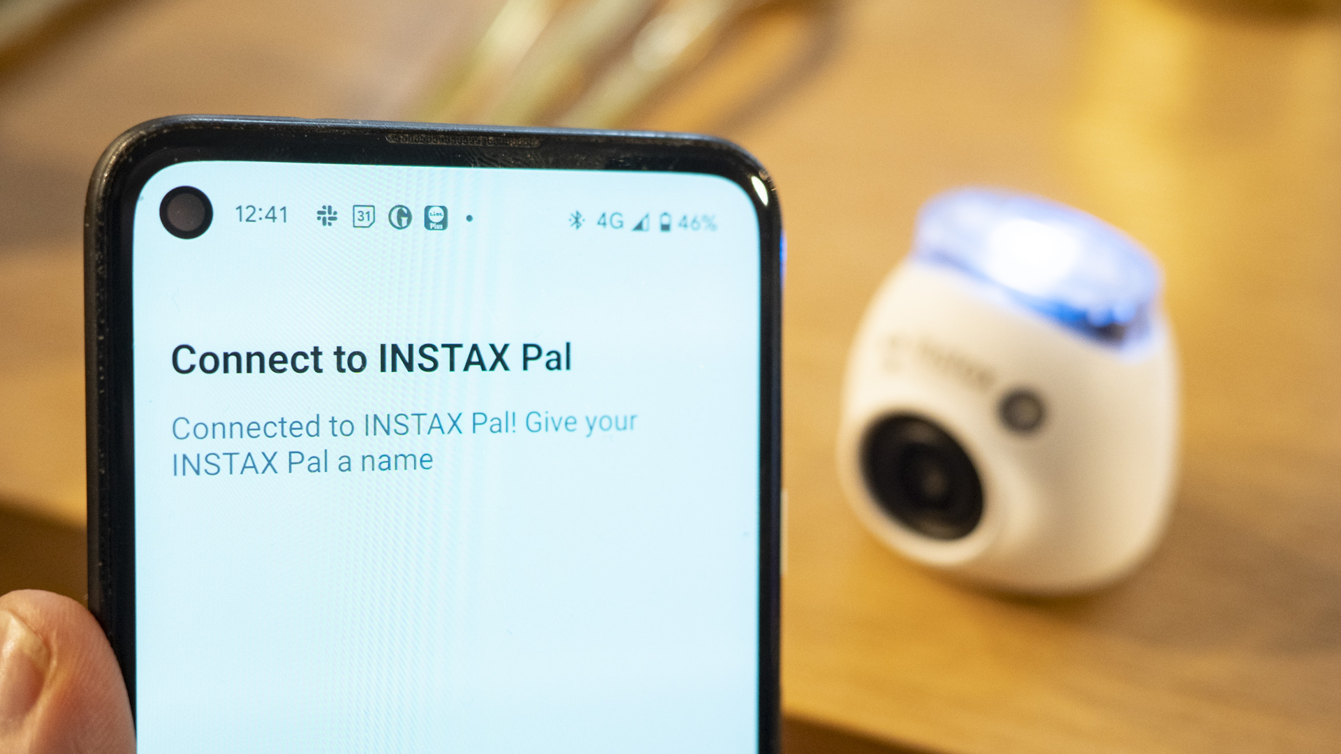 Instax Pal app device pairing process on phone display, with white Fujifilm Instax Pal in the background