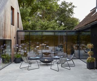 A concrete patio in front of a contemporary glass extension with table and chairs and a firepit