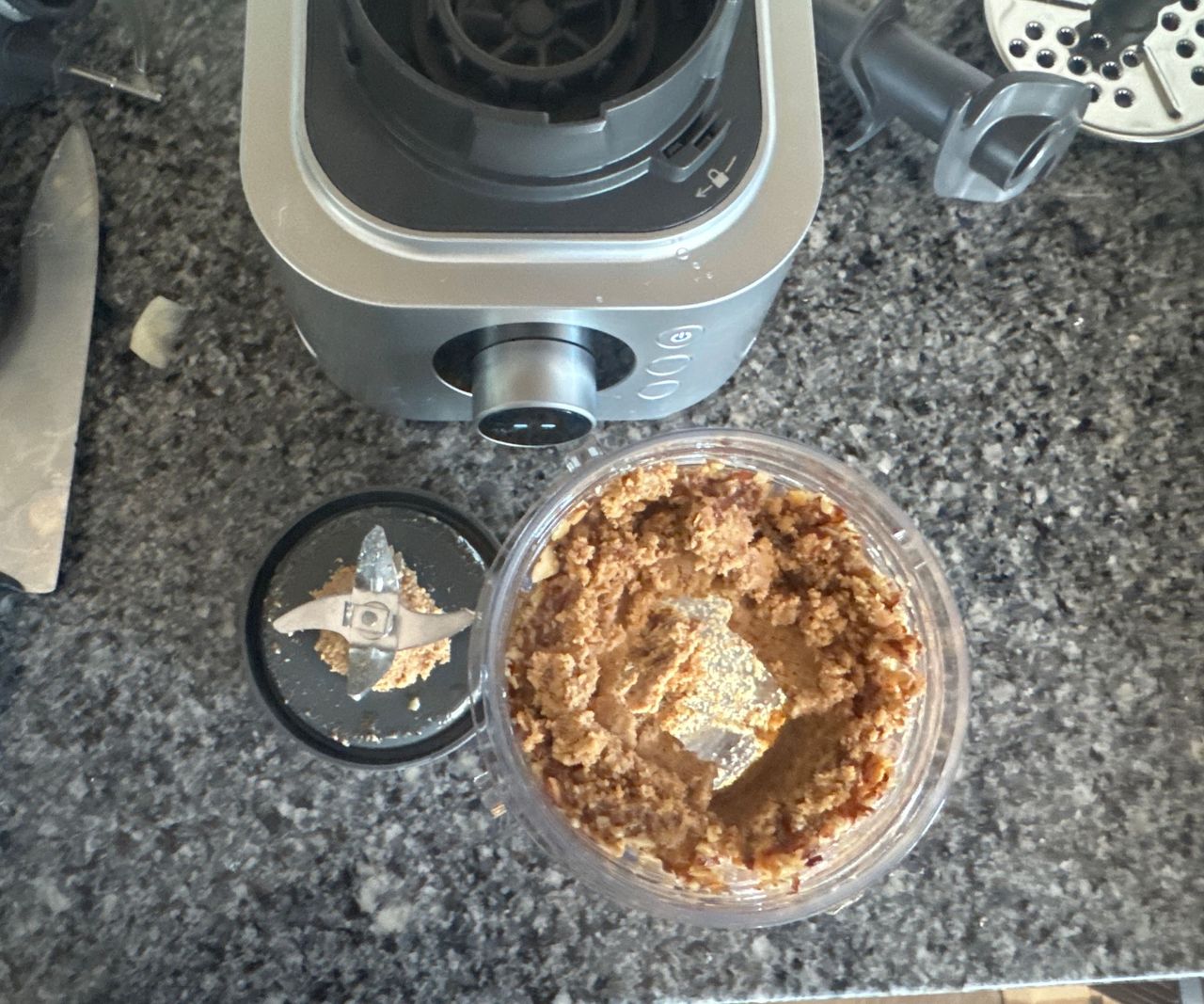 A top-down view of blended nut butter in a food processor cup beside the blender base.