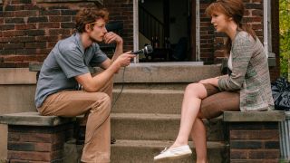 Mike Faist interviews Jodie Comer with a microphone while sitting on a stoop in The Bikeriders.