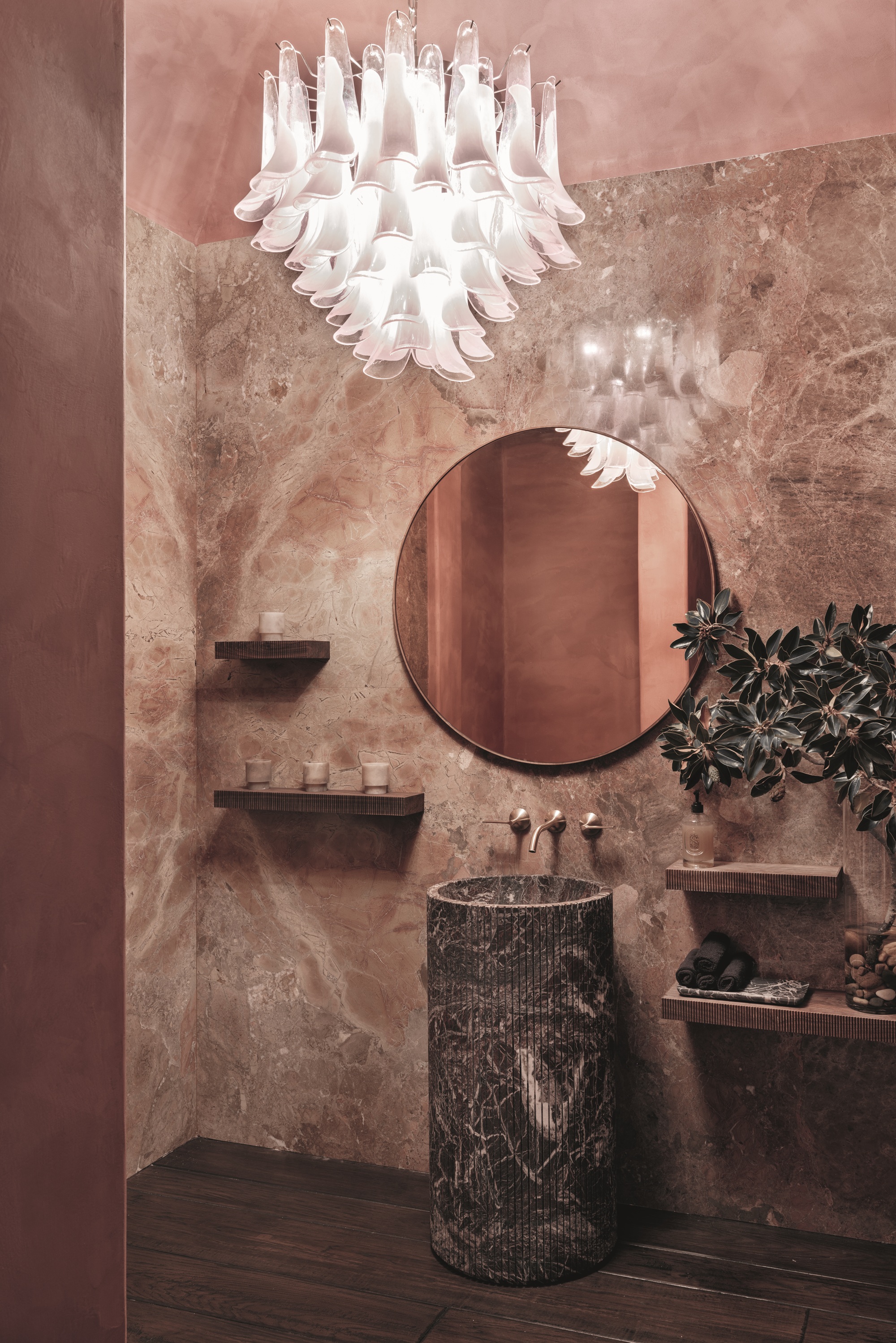 powder room with pink marble walls, pink plaster ceiling, fluted marble pedestal sink, round mirror, custom chandelier, and dark timber floors, with floating shelves on wall
