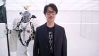 Hideo Kojima in a black blazer and t-shirt stands in a white room next to a person wearing a futuristic white armored suit holding a black flag.