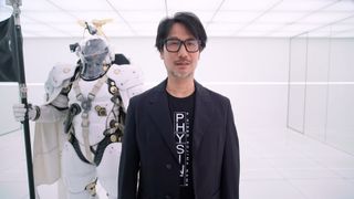 Hideo Kojima in a black blazer and t-shirt stands in a white room next to a person wearing a futuristic white armored suit holding a black flag.