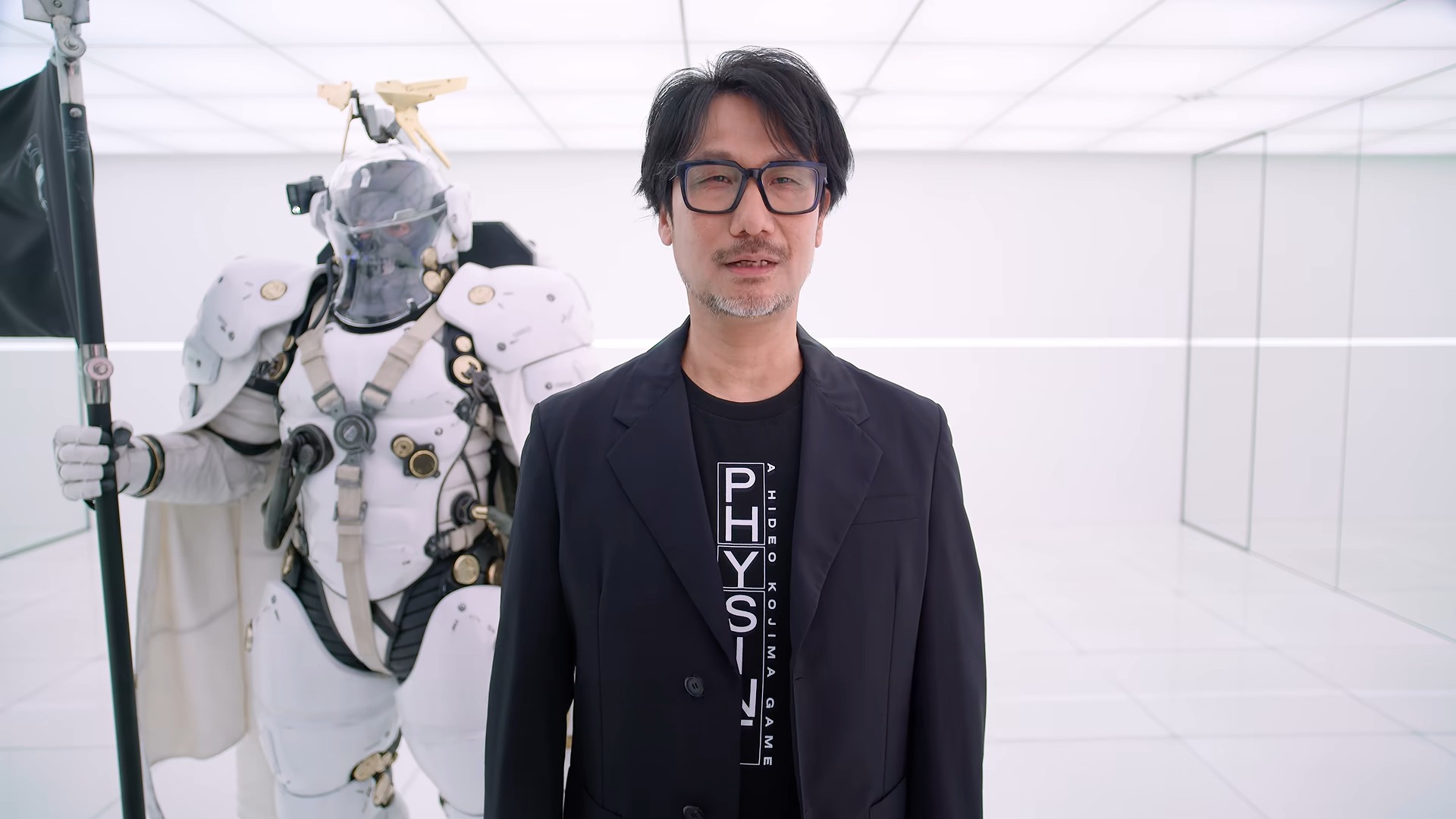 Hideo Kojima in a black blazer and t-shirt stands in a white room next to a person wearing a futuristic white armored suit holding a black flag.