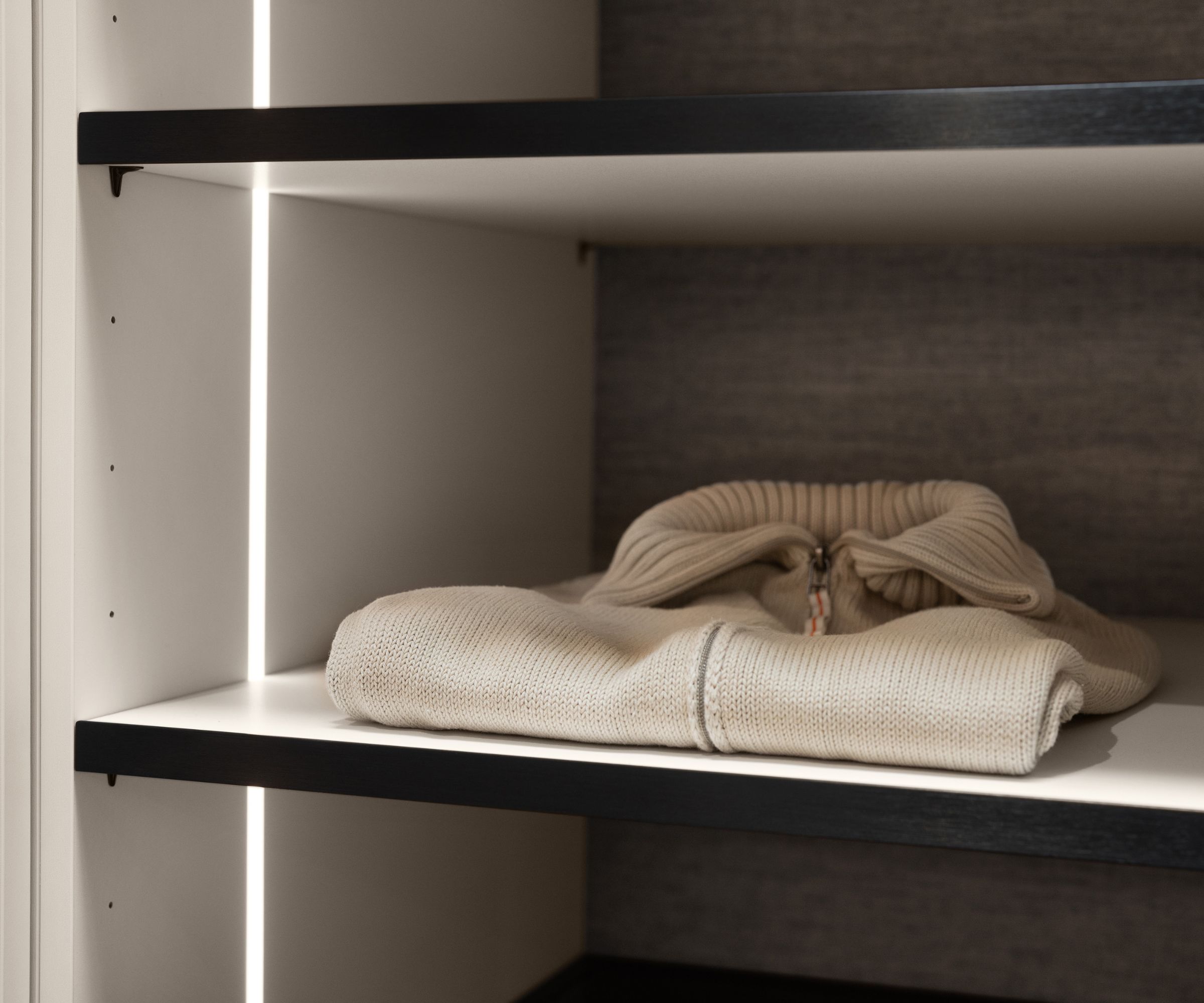 A close-up view of a white wardrobe shelf, neatly displaying a folded beige knitted sweater. Slim vertical LED strip lights line the edges of the unit, casting a warm glow across the fabric.