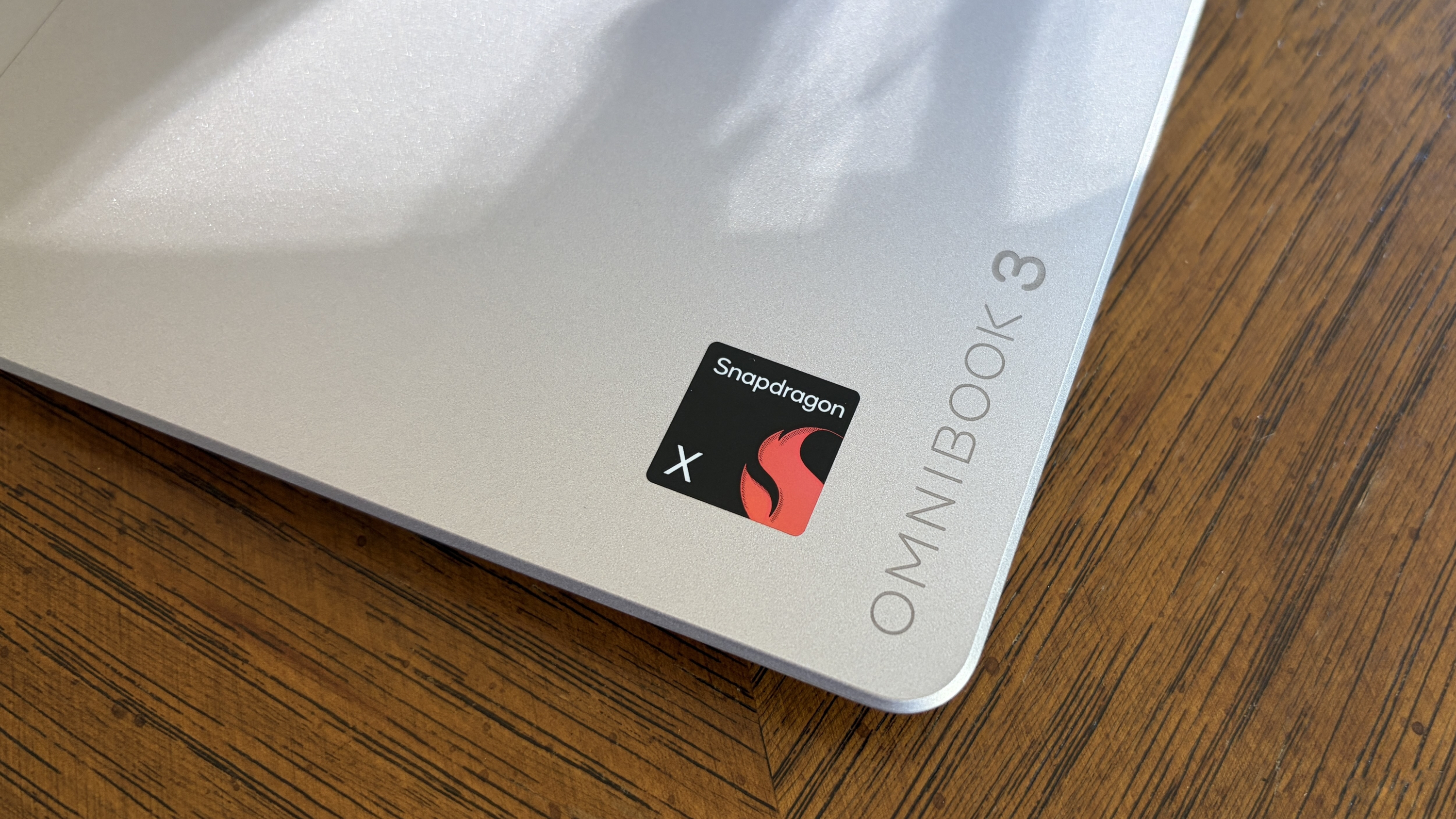 Close-up of a silver laptop lid labeled "OMNIBOOK 3" with a black Snapdragon sticker featuring a red flame logo on a wooden table.
