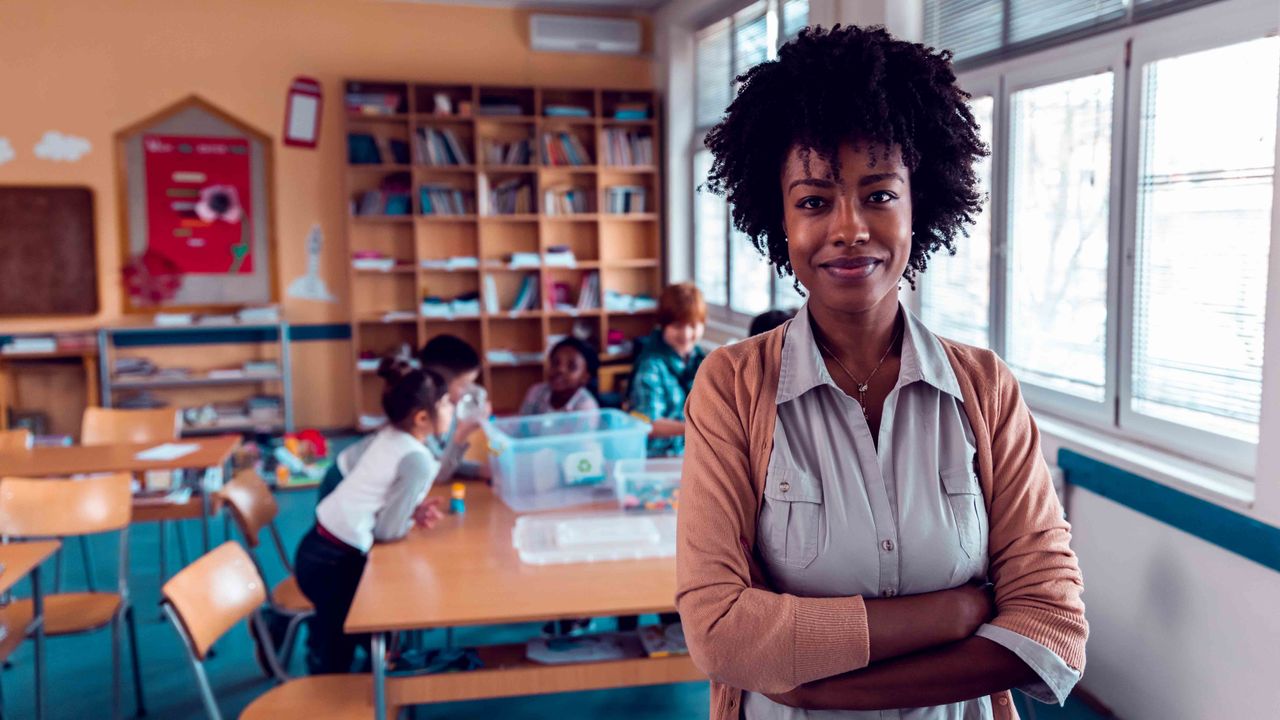 black woman with curly hair, arms crossed standing in front of elementary students