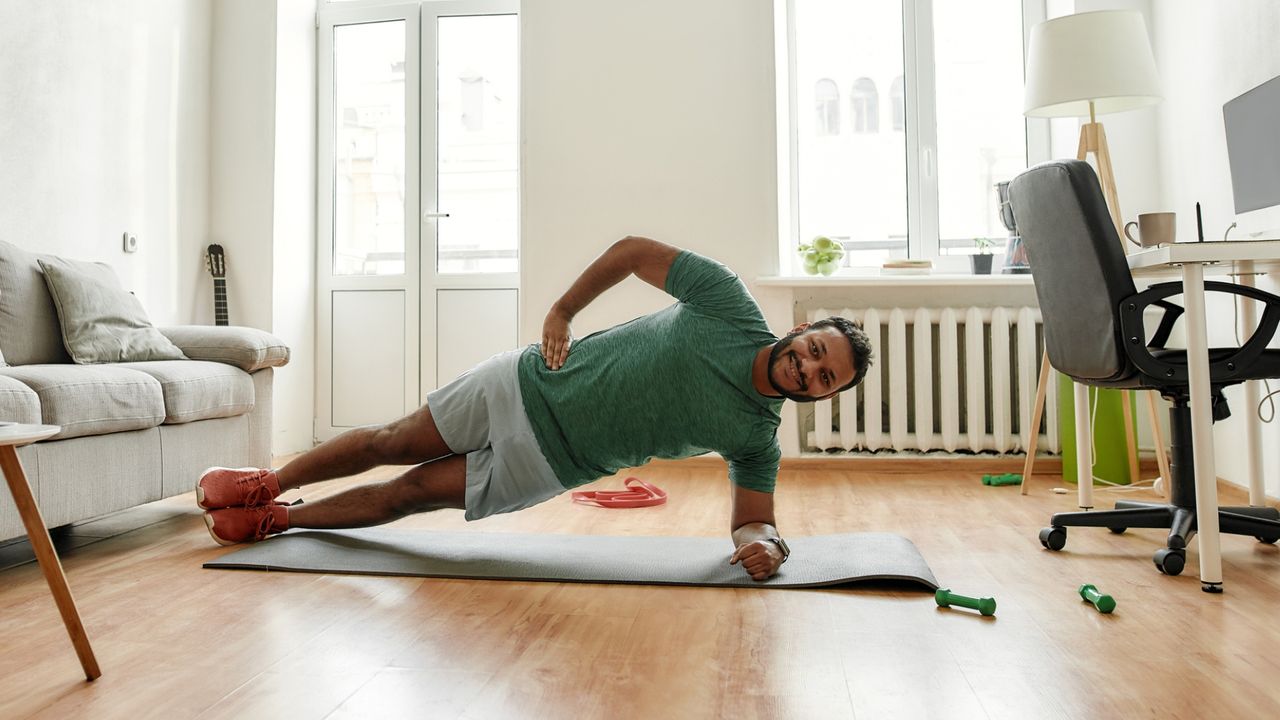 man in a living room setting doing a side plank facing the camera on a yoga mat, wearing a green tshirt and light shorts. 