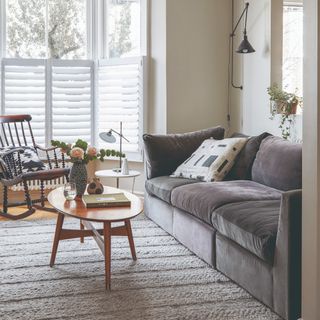 A small living room with a sectional grey velvet sofa on a textured rug and with an oval mid-century modern coffee table