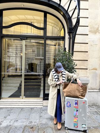 a woman standing outside a building next to her suitcase and tote wearing a baseball cap, blue jeans, and a tan trench coat with a striped sweater draped over her shoulders