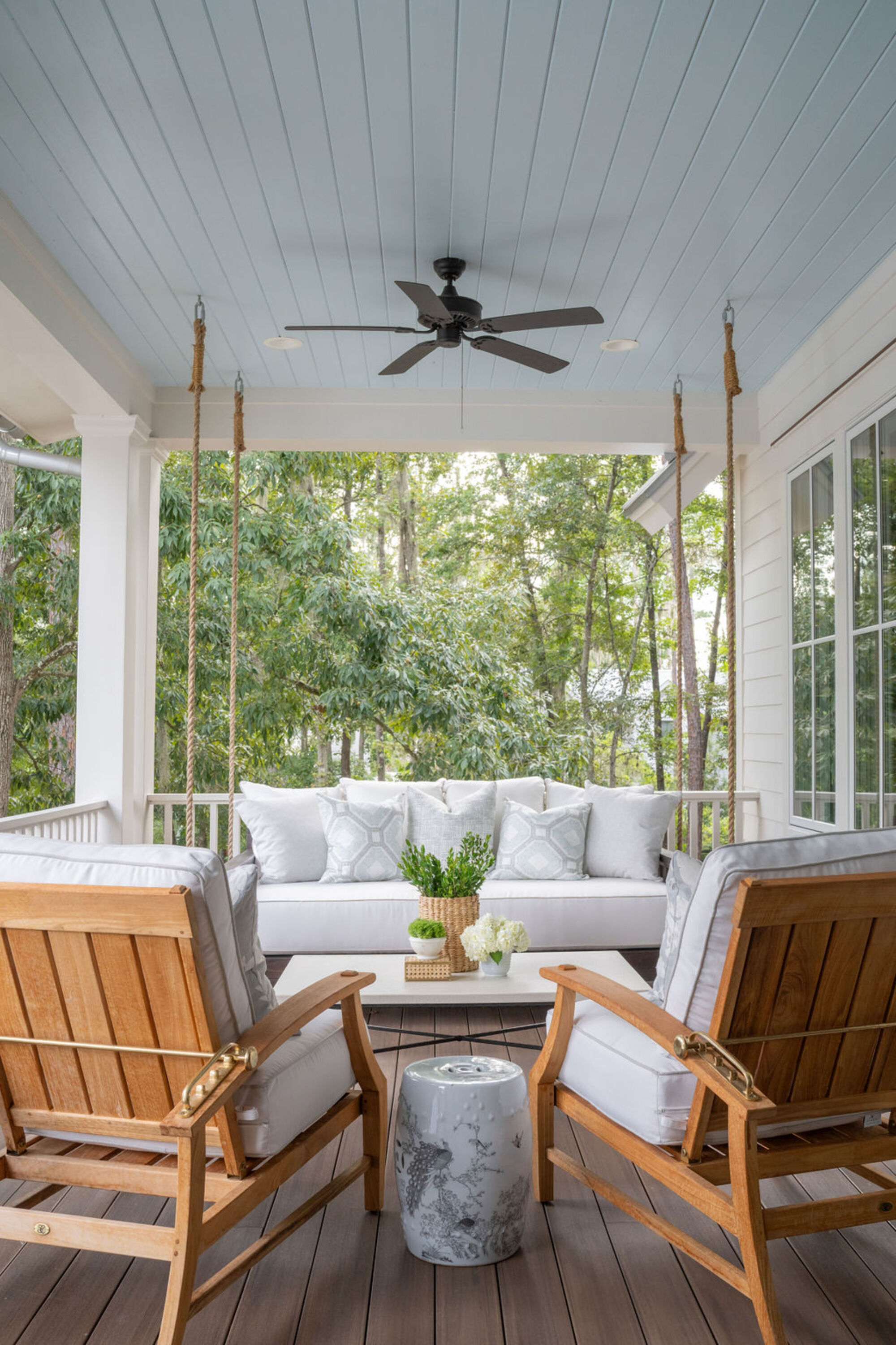 small outdoor area on deck with blue panelled ceiling with black ceiling fan, outdoor swing chair with white cushions, two outdoor chairs with white cushions, and cement side table, and a rectangle coffee table between the seating