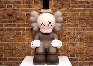 A sculpture by the artist KAWS with a skull sitting on a white platform in front of a brick wall