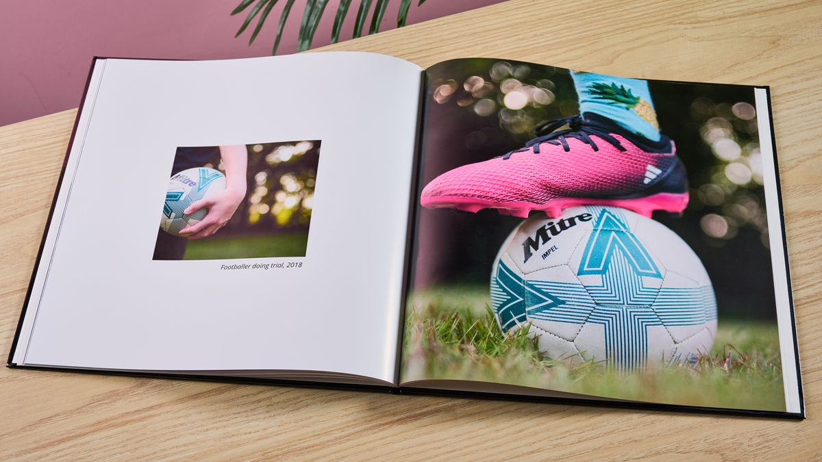 Vista Print photo book open on pages with photos of football and footballer, on table with pink wall and plant in background