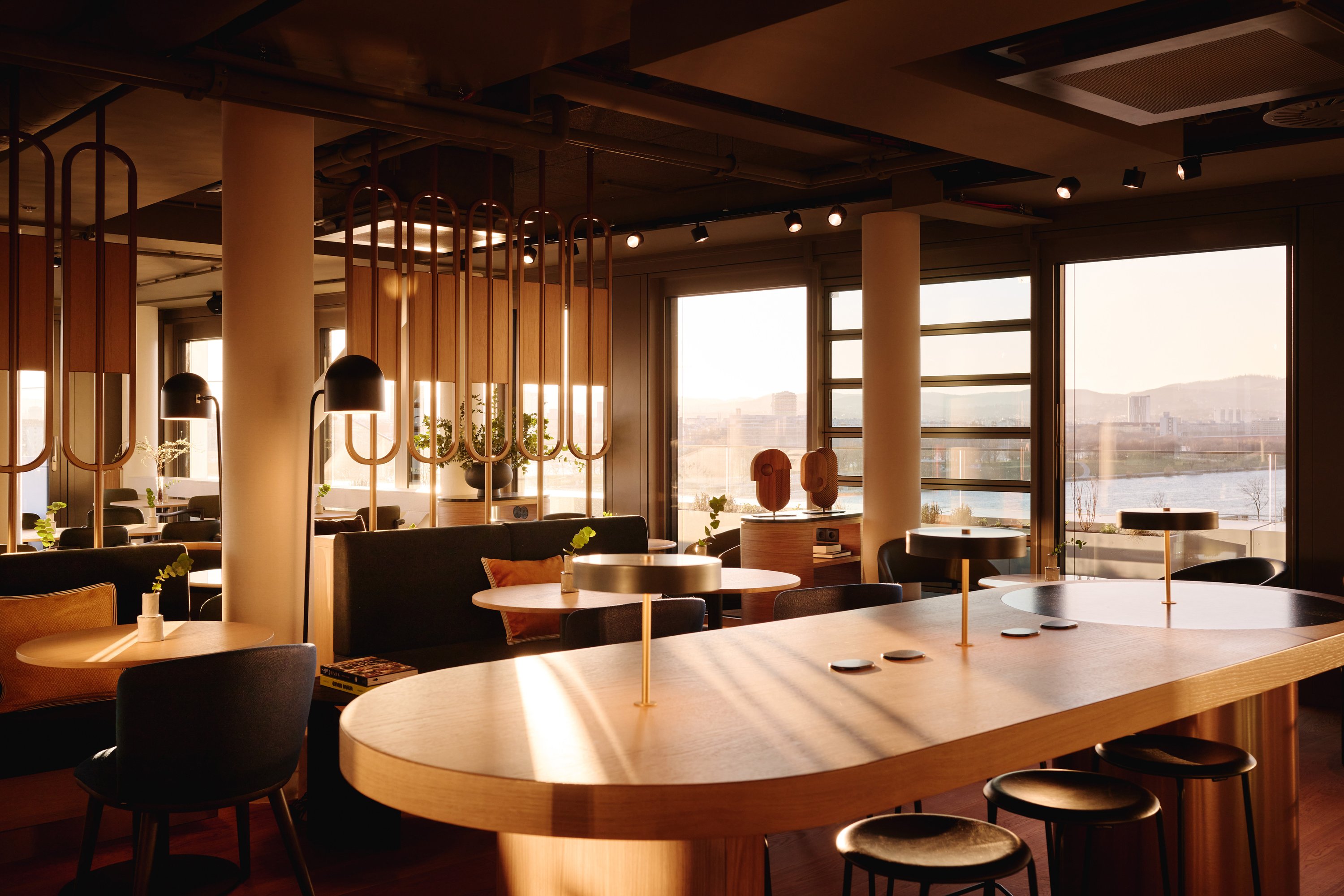 A sun-filled contemporary hotel boasts floor-to-ceiling windows, soft wooden tones mixed with earthy or green shades, mid-century modern-style furniture, and colorful collectibles all around.