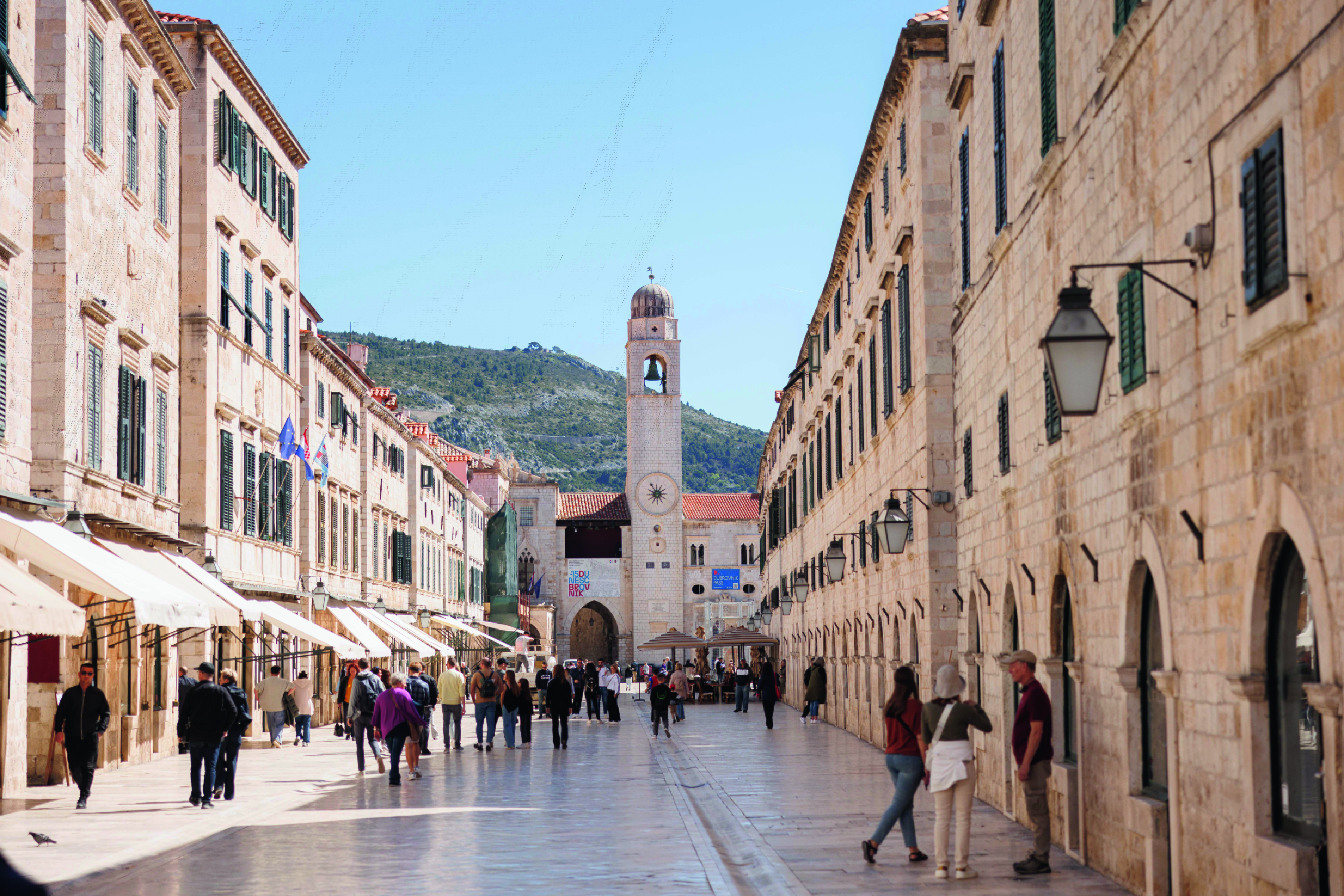 A wide street in Dubrovnik's old town, with cream brick buildings on either side and a church tower in the distance. 