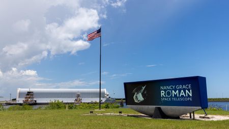 a long barge in the background arrives at a small offloading dock. In the foreground, and American flag in the center, and a digital display that says, "Nancy Grace Roman Space Telescope".