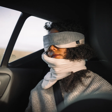 A woman resting in a car using a Trtl neck pillow and eye mask.