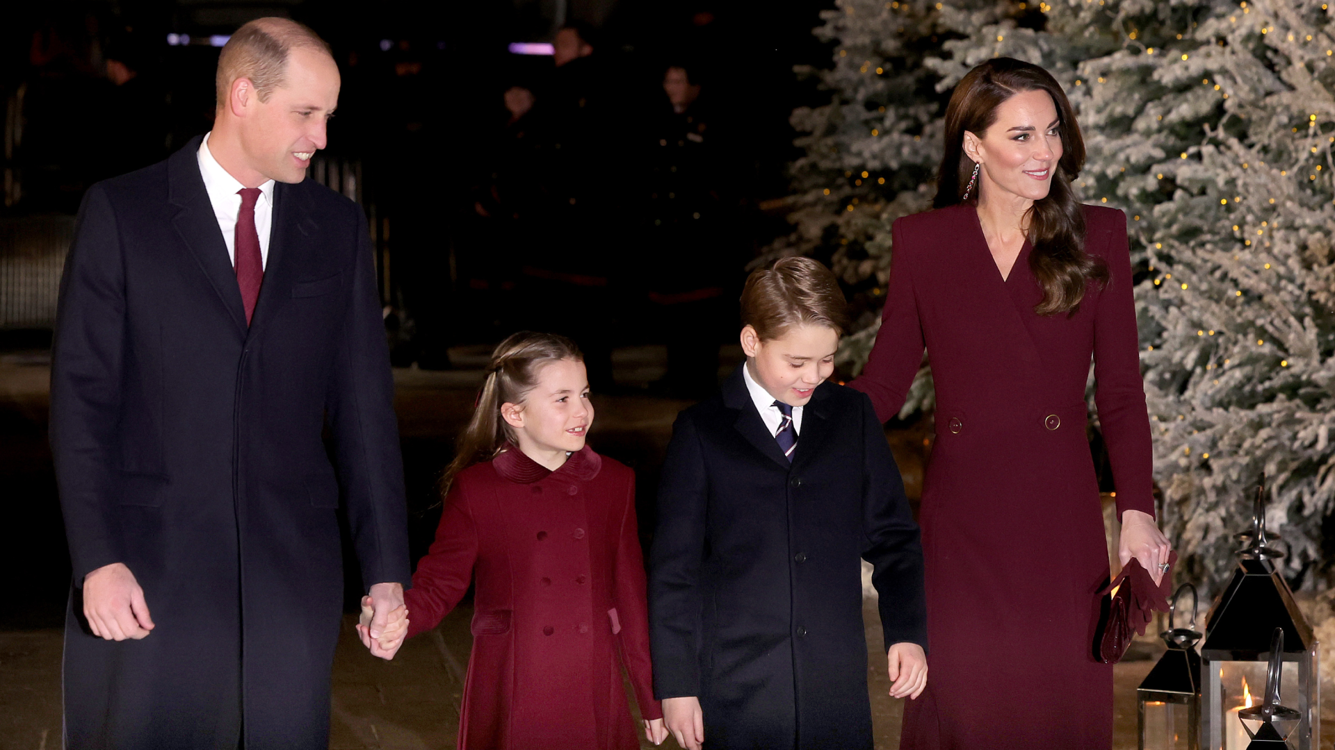 Princess Charlotte wearing a burgundy coat and smiling holding Prince William&#039;s hand, walking with Prince George and Princess Kate next to a snow covered tree