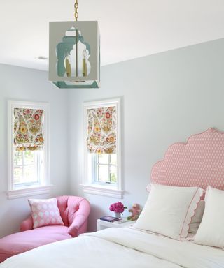 Bright, light bedroom with blue painted walls, two small windows with patterned roman blinds, double bed with large patterned headboard, pink chaise underneath window with scatter cushion