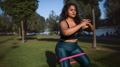 A woman in a sports bra and leggings is exercising in a tree-filled park on a sunny day. She is in a half-squat position, with a resistance band looped round her thighs and her knees gently bent. Her hands are clasped in front of her.