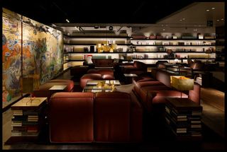 Interior view of Tsutaya Books featuring wood flooring, dark coloured leather seating, side tables made up of stacked books, lamps, wall art and wall-mounted shelving filled with items