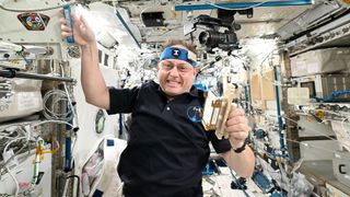 a man in a black short sleeve shirt and a blue headband floats inside a space station module cluttered with science equipment