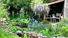 A garden featuring vegetables, shrubs., perennials and wisteria flowering over a pergola