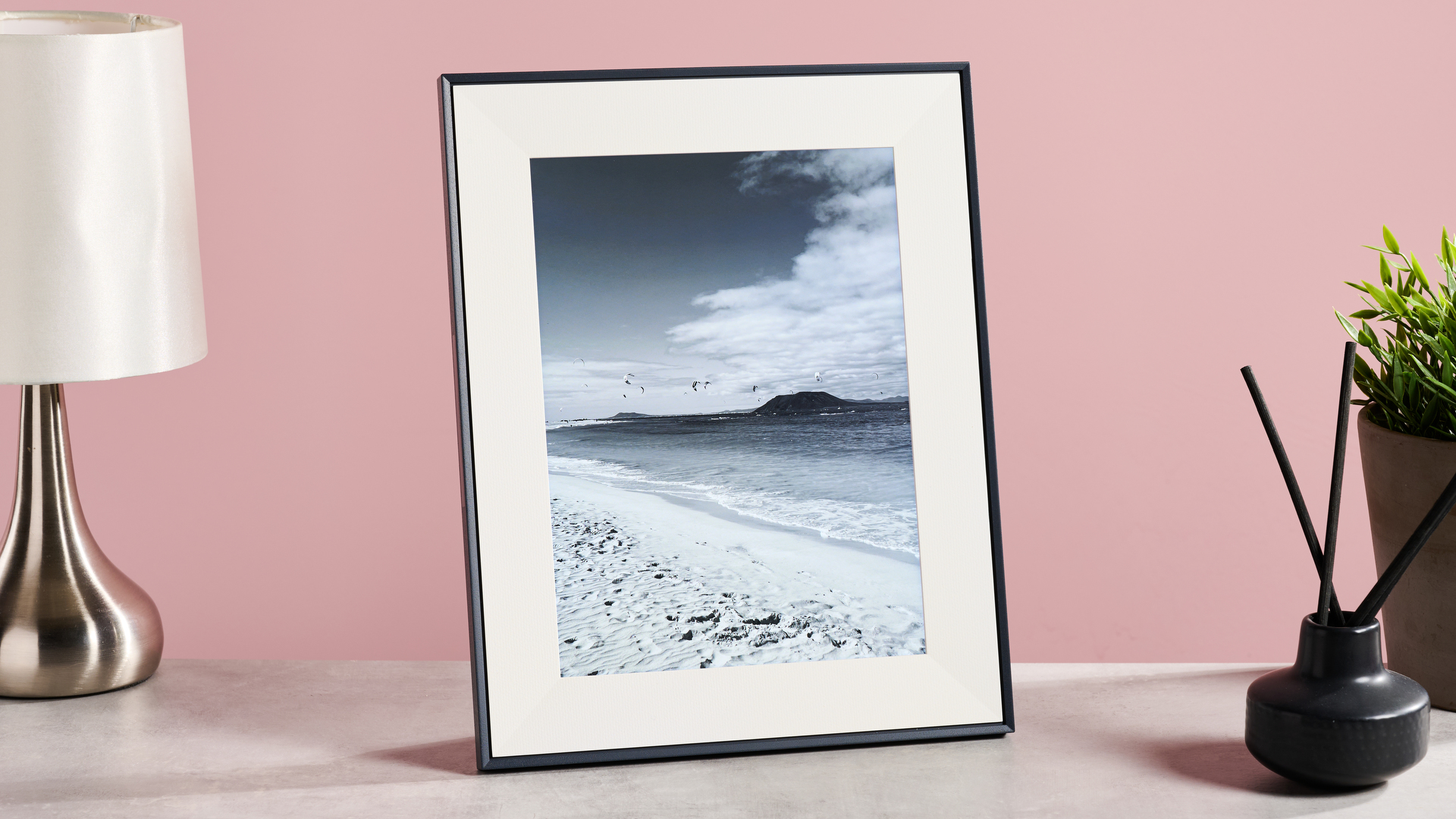 The Aura Aspen with a black and white photo of a beach and windsurfers, on a table with a lamp, reed diffuser and plant in front of a pink background.