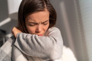 A young adult coughing into her arm.