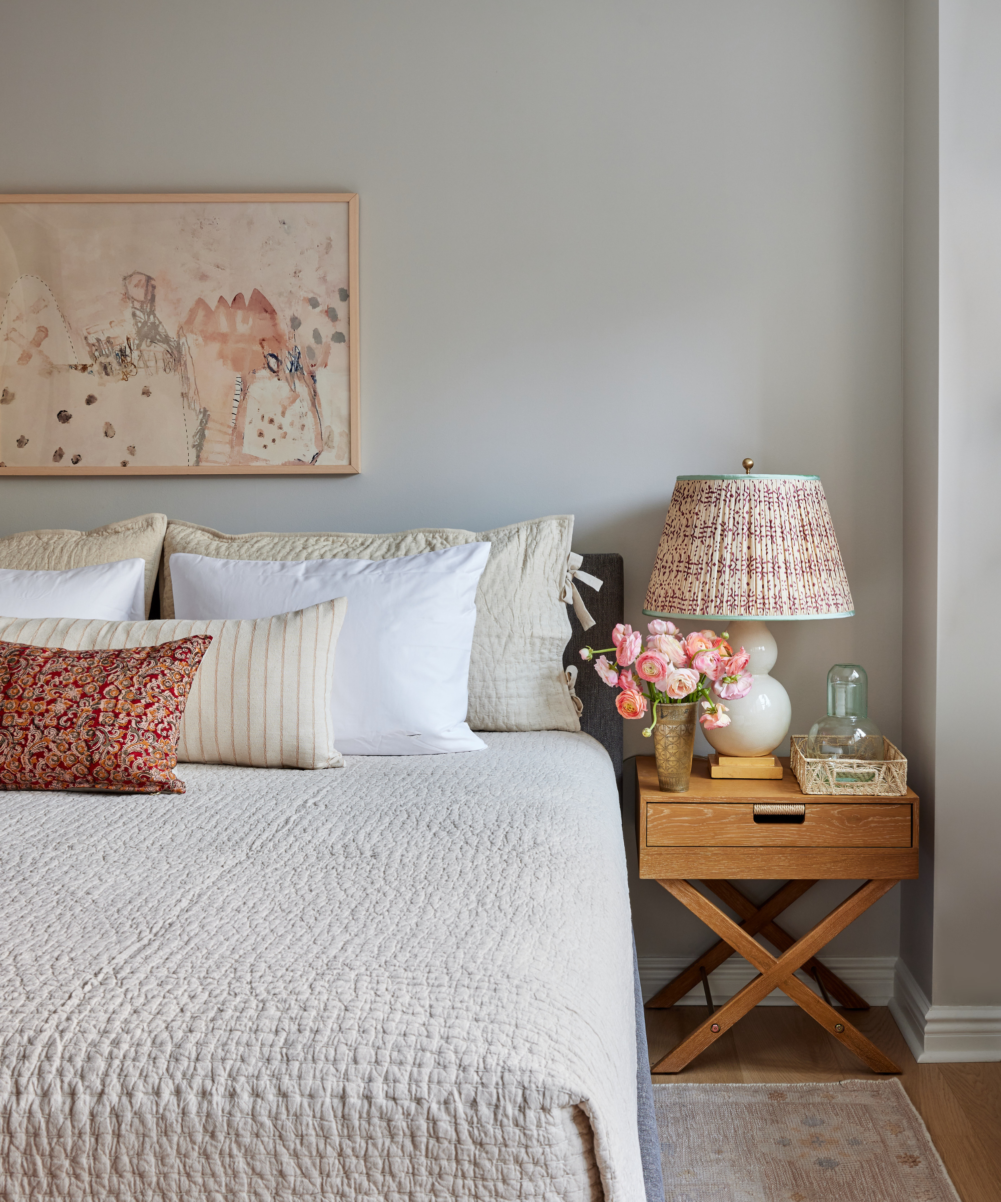 A greige bedroom with a textured bedspread, pink patterned lamp, and a wooden bedside table