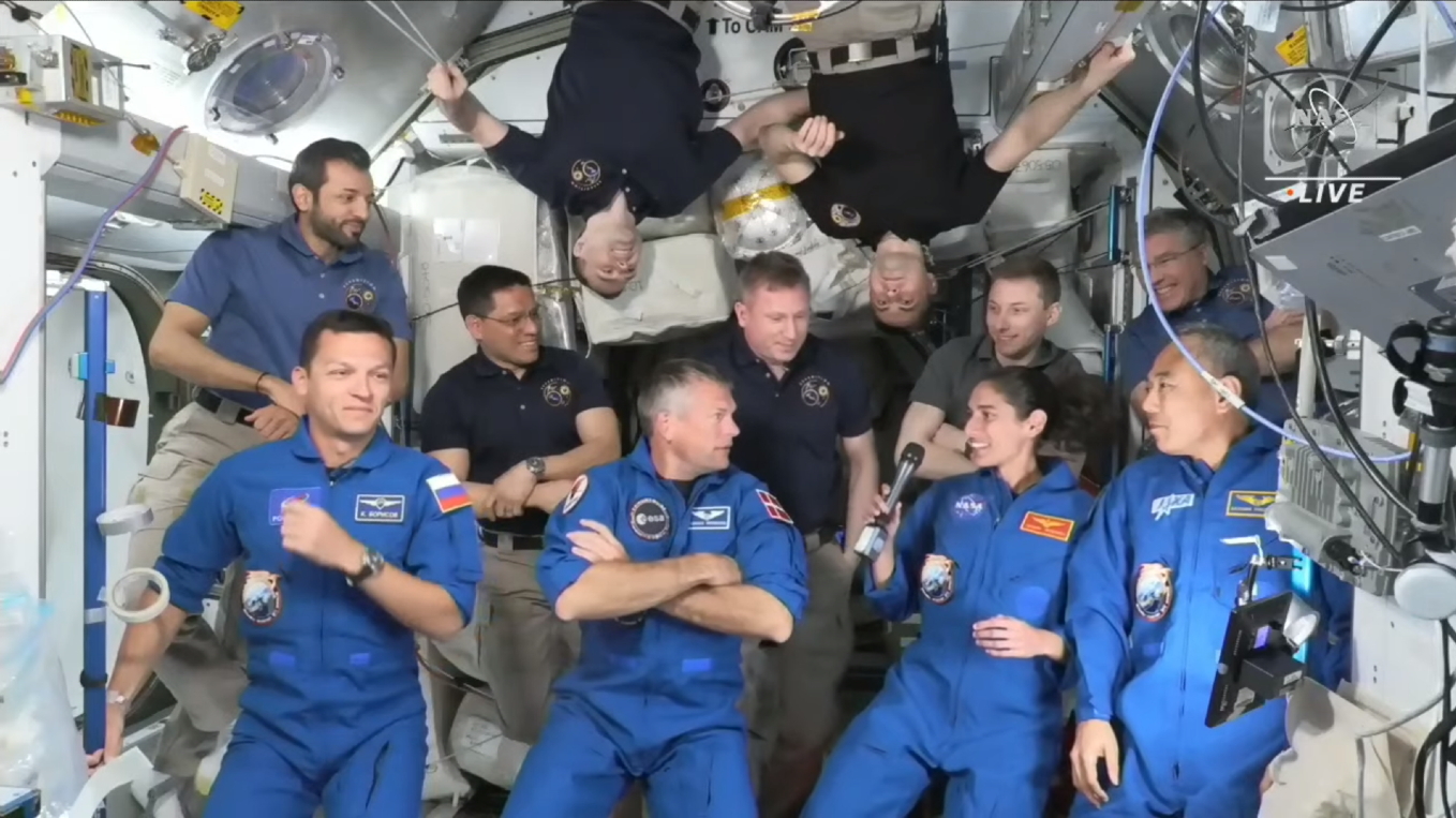 Top Stories Tamfitronics Eleven astronauts in different color uniforms gather on a space station, two float upside down.