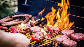 Flames flaring on a charcoal grill, cooking kebabs, burgers, and sausages