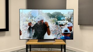 A 65-inch LG C4 OLED TV on a wooden table in the corner of a room. On the walls behind it are three large squares, and on screen is a man in a white baseball cap spraying a bottle of Ferrari-branded champagne with a crowd behind him.