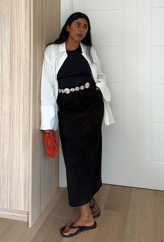 a summer wardrobe essential shown on a woman wearing minimal sandals with a white button-down shirt layered over a black tank top with a silver belt, a black maxi skirt, and a red bag