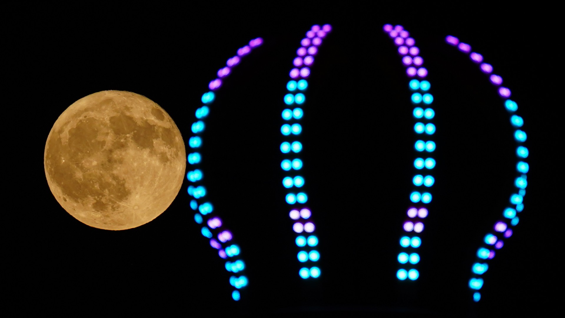 An orange full moon is pictured against a dark night sky. Dark lunar seas and prominent craters can be seen marking the lunar surface, while the neon lights adorning the tip of a carousel ride dominate the right-hand side of the picture.