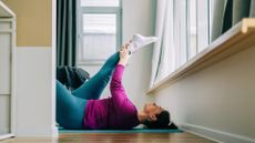 woman lying on her back stretching her leg upwards. she's wearing a cerise pink top and blue leggings on wooden floors indoors sideways to the camera under a window.