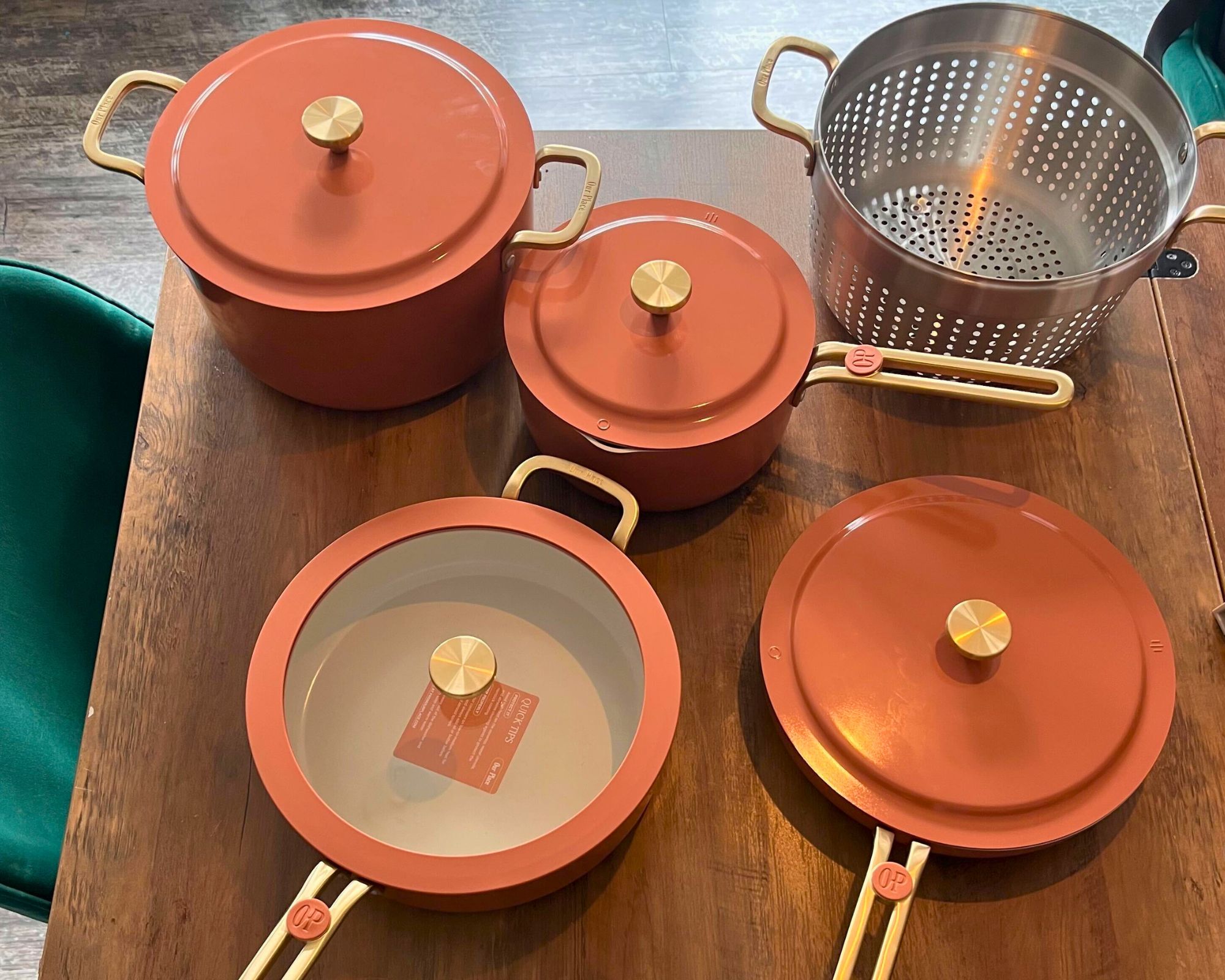 The Our Place 11-Piece Essentials Cookware Set on a Dining Table in Terracotta: It includes two wide shallow pans with lids (one has a glass window), a stock pot, a saucepan, and a stainless steel strainer