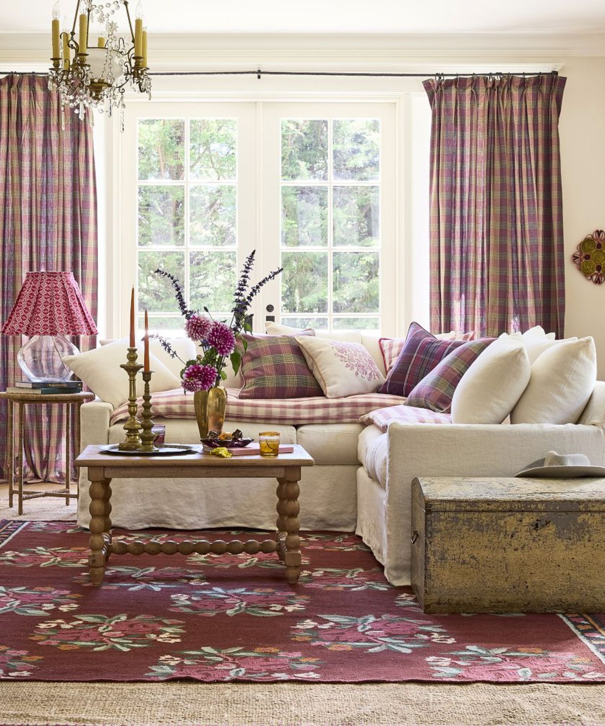 neutral living room with large french doors and a white corner couch styled with pink plaid pillows with matching curtains, vintage style wooden furniture and a floral flatweave rug