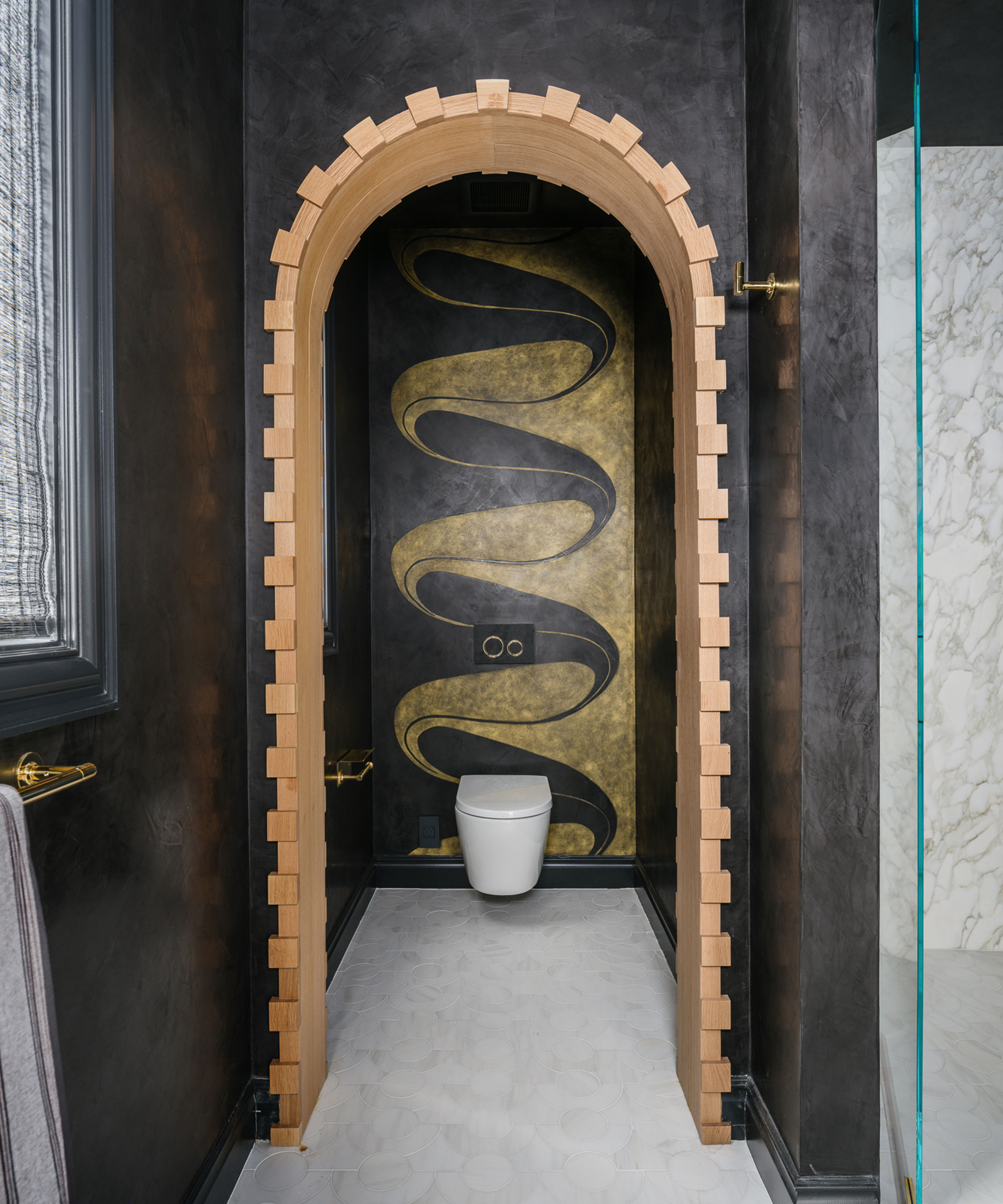 A modern bathroom featuring a notched light wood archway leading to a toilet. The back wall has a bold gold and black wavy mural, complemented by dark textured walls and white circular floor tiles.