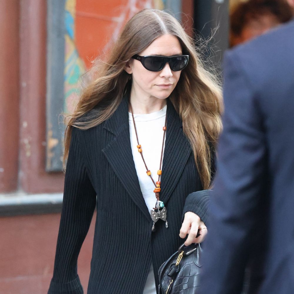 Ashley Olsen wears a black pleated blazer over a white t-shirt with black pleated trousers.