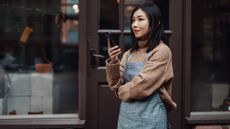 Young woman wearing an apron speaking on her phone, standing in front of a small business or shop that she owns