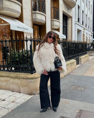 Ellie wears a white fur coat, black mini bag, black wide-leg pants, and black heels.