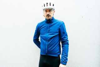 A white man in a royal blue winter cycling jacket stands against a white wall