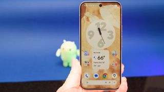 Google Pixel 10 Pro XL in Porcelain against a blue background with a green neon Android figurine in the background